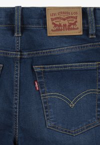 Jeans de mezclilla azul oscuro con una etiqueta de cuero, costuras amarillas y un diseño de bolsillo curvo distintivo. Etiqueta roja de Levi's en el bolsillo trasero.