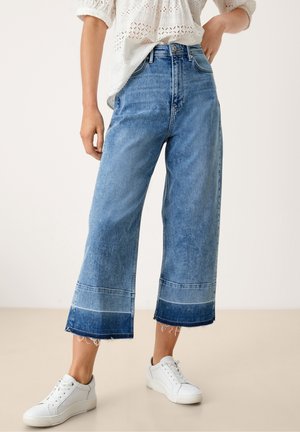 Donna che indossa jeans larghi a gamba corta di colore azzurro chiaro con orli sfrangiati, camicetta bianca con occhielli e sneakers bianche, in piedi all'interno.