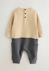 Beżowy i szary dwuczęściowy zestaw. Bluza z trzema czarnymi guzikami z tyłu, szare spodnie z bocznymi kieszeniami i ściągaczami.