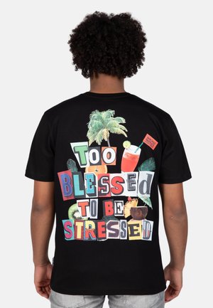 Zwart T-shirt met een kleurrijk grafisch ontwerp met de tekst "Te Gelukkig Om Gestrest Te Zijn" naast tropische elementen en drankjes.