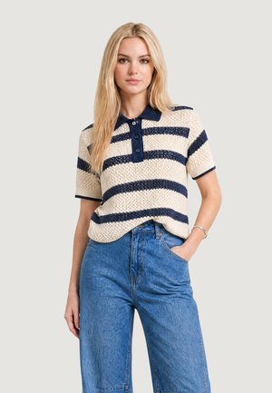 Femme aux longs cheveux blonds portant un polo en tricot à manches courtes rayé crème et bleu marine et un jean bleu taille haute, main dans la poche.