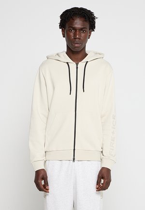 Young man wearing a cream zip-up hoodie with black drawstrings, front pockets, and light gray sweatpants, standing against a plain background.