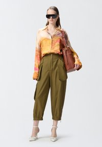 Šarena košulja s dugim rukavima i dugmadi; maslinasto zelene hlače širokog kroja s bočnim džepovima; smeđa strukturirana torba; bijele cipele na visokoj peti.