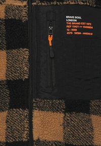 Casaco de fleece xadrez em castanho e preto, com um bolso com fecho éclair com um puxador laranja e marcação em texto branco e laranja.