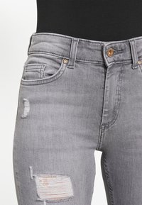 Jeans em denim cinza com cintura alta, textura desbotada e detalhes desgastados com fios expostos e fechamento clássico por botão.