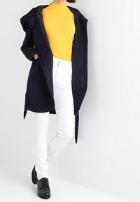 Marinblå hooded kappa över en gul ribbad polotröja, i kombination med vita skinny jeans och svarta skor. Kappan har en mjuk textur och knappar.