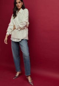 Pull crème avec un motif de losanges texturé et des embellissements, superposé sur une chemise blanche. Jean droit bleu clair et escarpins argentés.