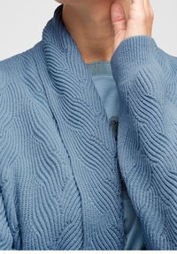 Cardigan lavorato a maglia blu chiaro con un motivo a onda testurizzato. Presenta un ampio colletto e maniche lunghe, indossato sopra una maglietta blu chiaro.