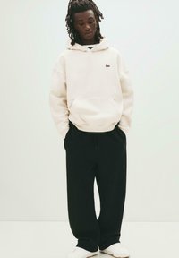 Person, der einen cremefarbenen Hoodie mit kleinem Logo, schwarze lockere Hosen und weiße Turnschuhe trägt, steht vor einem einfarbigen, hellen Hintergrund.