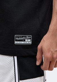 Μαύρο πουκάμισο Mitchell & Ness με λευκό ορθογώνιο λογότυπο κοντά στη ραφή και ένα χέρι που ακουμπάει δίπλα σε μαύρα και λευκά δίχτυα σορτς.