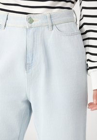 Světle modré denimové džíny s vysokým pasem, vybavené zapínáním na knoflík, poutky na opasek a předními kapsami. Hladká textura a minimální detaily šití.