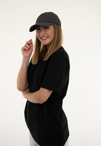 Čierne oversized tričko vyrobené z bavlny, skombinované s tmavosivou baseballovou čiapkou. Pozadie je jednofarebné biele. Model stojí v hravej póze.