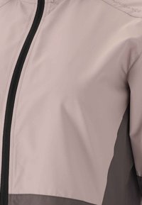 Lys grå vindjakke har en sort lynlås, en glat overflade og et kontrastende mørkegrå panel i bunden. Branding er synlig på skulderen.