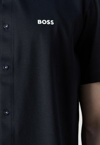 Zwarte korte mouwen shirt van een gladde stof, met een klassieke kraag, knoopsluiting en wit "BOSS" logo boven het linkerborstgebied.