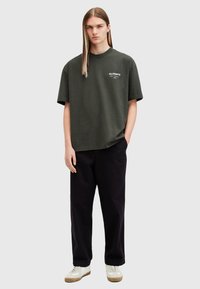 Camiseta de gran tamaño en verde oscuro con cuello redondo y estampado de logo en blanco en el pecho, combinada con pantalones negros y zapatillas blancas.