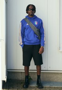 Μπλε φούτερ με κουκούλα και κίτρινες ρίγες, σήμα της Ιταλίας, μαύρα σορτς, μαύρες μποτάκια έως τον αστραγάλo και γυαλιά ηλίου; στέκεται σε έναν λευκό τοίχο.