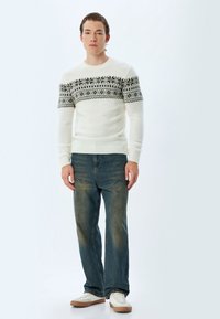 Weißer Strickpullover mit grauen geometrischen Mustern auf der Brust, kombiniert mit verwitterten blauen Jeans und weißen Sneakers mit braunen Sohlen.