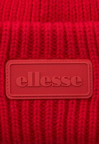 Červená pletená látka se čtvercovou červenou gumovou nášivkou s vyraženým názvem značky "ellesse" tučnými písmeny.