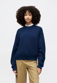 Sweater azul-marinho com textura canelada, decote redondo e fendas laterais. Mangas longas e largas com punhos justos. Estilizado sobre uma camisa clara.