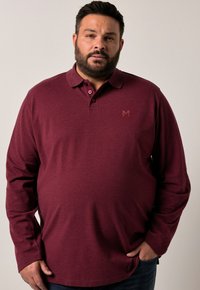 Muž s vousy, oblečený v burgundském polo tričku s dlouhým rukávem a knoflíky, a tmavých džínách, stojící před jednolitým světlým pozadím.