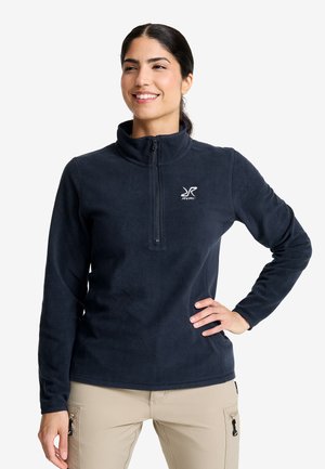 Frau mit dunklem Haar, die in einem schwarzen Fleece-Pullover und beigen Hosen lächelt, steht mit einer Hand auf der Hüfte vor einem weißen Hintergrund.