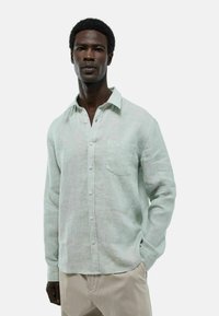 Chemise en lin vert clair avec col boutonné, poche frontale et manches longues. Tissu texturé avec une coupe décontractée.