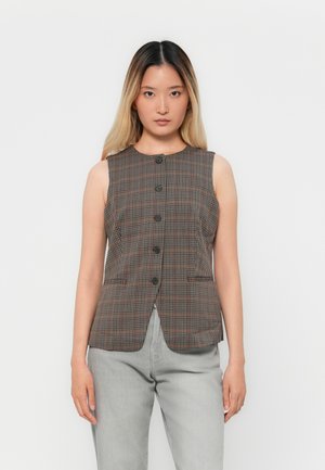 Woman with long ombre hair wearing sleeveless brown plaid buttoned vest and light gray pants, standing against plain white background.