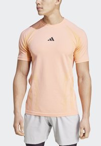 Camiseta deportiva corta de color rosa claro con mangas cortas, que presenta acentos texturizados en los laterales y un logo negro de Adidas en el pecho.