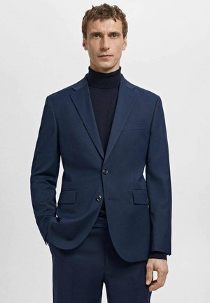 Navy textured blazer with two buttons, notch lapels, and two front pockets, worn over a black turtleneck. Slim fit design.