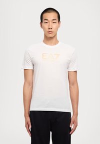 Hvit bomulls t-skjorte med en gul logo "EA7" på forsiden. Korte ermer og klassisk rund hals. Glatt tekstur, avslappet passform.