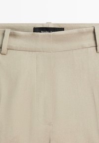 Béžové kalhoty na míru s poutky na opasek, skrytým předním zapínáním a viditelnou černou štítkem uvnitř pasu.