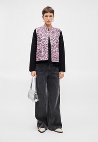 Ροζ λεοπάρ εκτύπωση γιλέκο πάνω από ένα μαύρο μακρύ μανίκι τοπ, συνδυασμένο με μελανί φαρδιά παντελόνια και μια μικρή ασημένια τσάντα.