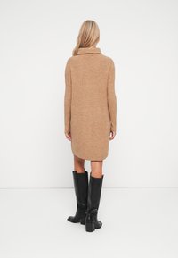 Vestito a maglia a maniche lunghe con colletto a imbuto in un tessuto marrone chiaro, lungo fino al ginocchio, abbinato a stivali alti neri con tacco, dalla texture liscia.
