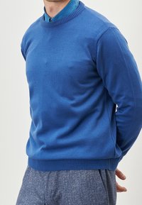 Maglione blu a girocollo realizzato in un tessuto a maglia liscia con rifiniture a coste. Indossato sopra una camicia blu con colletto, abbinato a pantaloni grigi dalla texture.