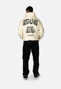Felpa crema con grande testo nero sul retro: "CRYSTALPARIS CHIAMAMI IN UNA NOTTE NORDICA D'ORO +00 (33)". Abbinata a pantaloni neri.
