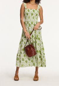Florales Maxikleid in Weiß mit grünen Designs, quadratischer Ausschnitt und Rüschenträgern; kombiniert mit einer braunen Bucket Bag und tanfarbenen Plateausandalen.