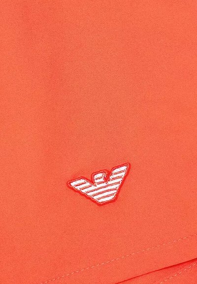 Un tejido naranja con una textura suave presenta un logo bordado en forma de pájaro con rayas rojas y blancas.