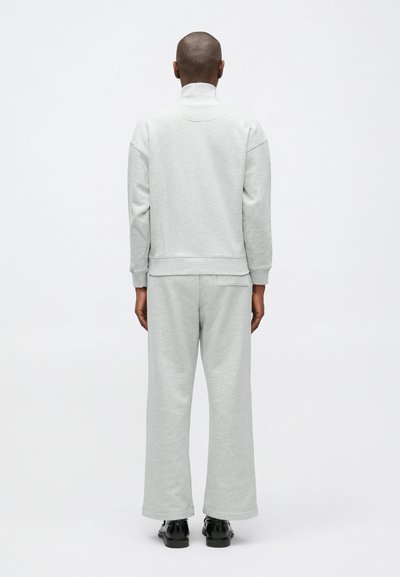 Osoba stojaca chrbtom, oblečená v svetlosivej mikine a širokých nohaviciach, s čiernymi topánkami na bielom pozadí.