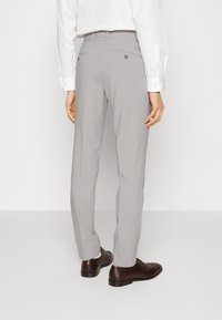 Pantaloni grigi su misura con una texture liscia, dotati di due tasche posteriori, indossati con scarpe brogue in pelle marrone. Camicia bianca visibile.