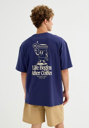 Persona con cabello rizado que lleva una camiseta azul marino con una taza de café de dibujos animados que dice "¡Hola!" y el texto "La vida comienza después del café" en la parte trasera.