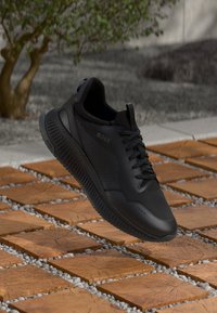 Mustad mustreeritud kangrega mustad spordijalatsid, millel on võrgupaneelid, tekstuuriga kummitald, silmapaistev logo ja lamedad paelad. Rippuvad mustritud telliskivipinna kohal.