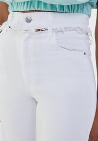 Pantalones vaqueros blancos de tiro alto con dobladillo desgastado y detalles de borde sin terminar, que incluyen un bolsillo frontal y herrajes de botón plateado. Textura de mezclilla suave.