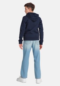 Ragazzo in jeans azzurri, sneakers bianche e felpa blu navy con cappuccio abbassato, in piedi con la schiena rivolta contro uno sfondo bianco.