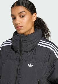 Een jonge persoon met golvend haar draagt een zwarte pufferjack met witte strepen en een Adidas-logo, en kijkt zelfverzekerd naar de kijker.