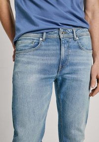 Vaqueros de mezclilla azul claro con un acabado desgastado, diseño de cinco bolsillos, herrajes plateados y costuras en contraste. Llevados con una camiseta azul.