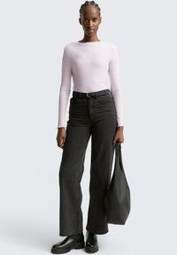 Vrouw met gevlochten haar, gekleed in een lichtroze geribbelde lange mouwen top, hoog getailleerde zwarte wijde broek, zwarte laarzen, en ze houdt een zwarte tote bag vast.