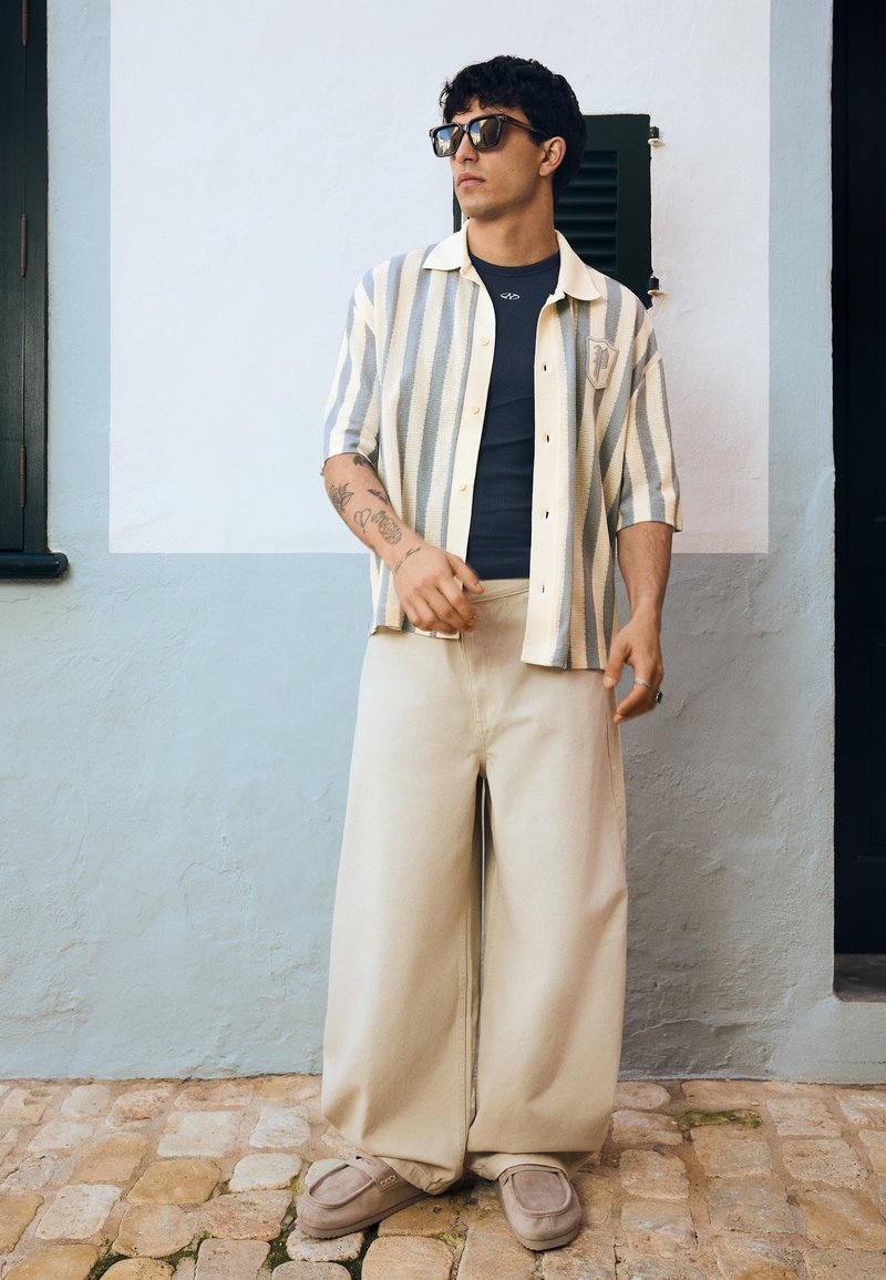 Férfi napszemüvegben, fekete top fölött csíkos rövid ujjú ing, széles bézs nadrág és mokaszin, macskaköveken áll egy fehér és világoskék fal mellett.