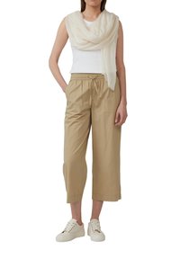 Beige cropped broek met een elastische tailleband, witte aansluitende tanktop en lichtcrème sjaal. Model draagt witte sneakers.