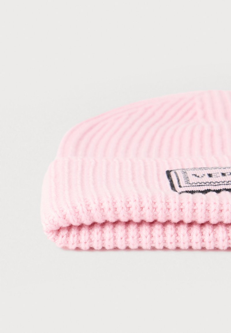 Pink knitted beanie with a ribbed texture, featuring a folded cuff and a rectangular label with black details sewn on the front.