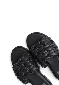 Sandales mules en cuir noir avec un dessus tressé, une texture douce et une semelle plate avec une zone de bout arrondi.
